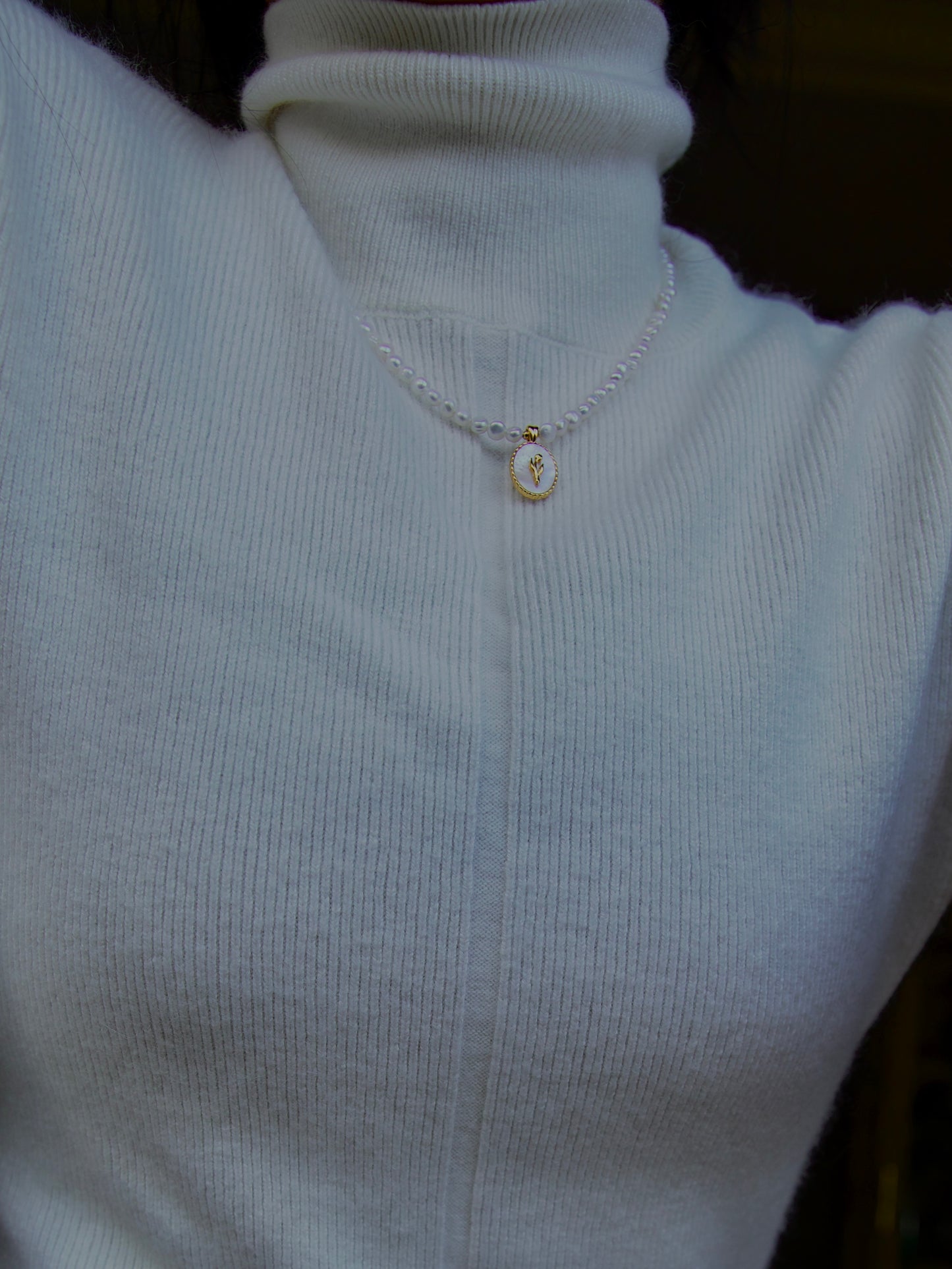 A close-up of a person wearing a white ribbed turtleneck sweater, showcasing a pearl necklace with an oval-shaped gold-plated pendant. The pendant features a mother of pearl base with a tulip design in gold at its centre. The necklace rests neatly around the neck, contrasting softly against the white fabric.